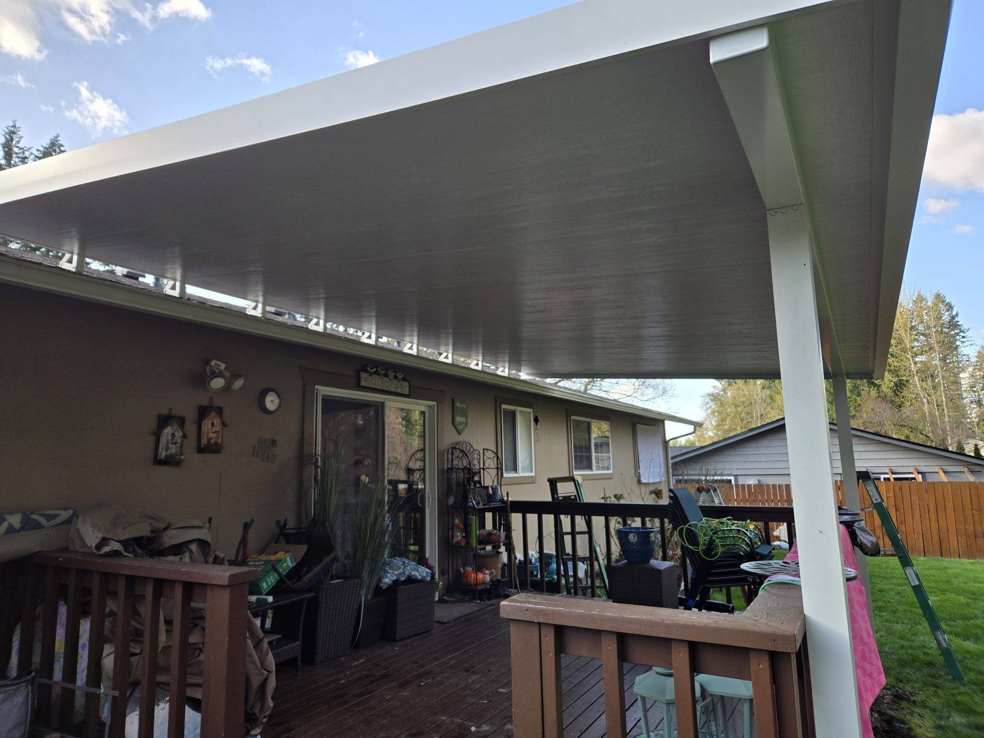 A patio featuring a covered porch and a wooden deck, surrounded by greenery and outdoor furniture.